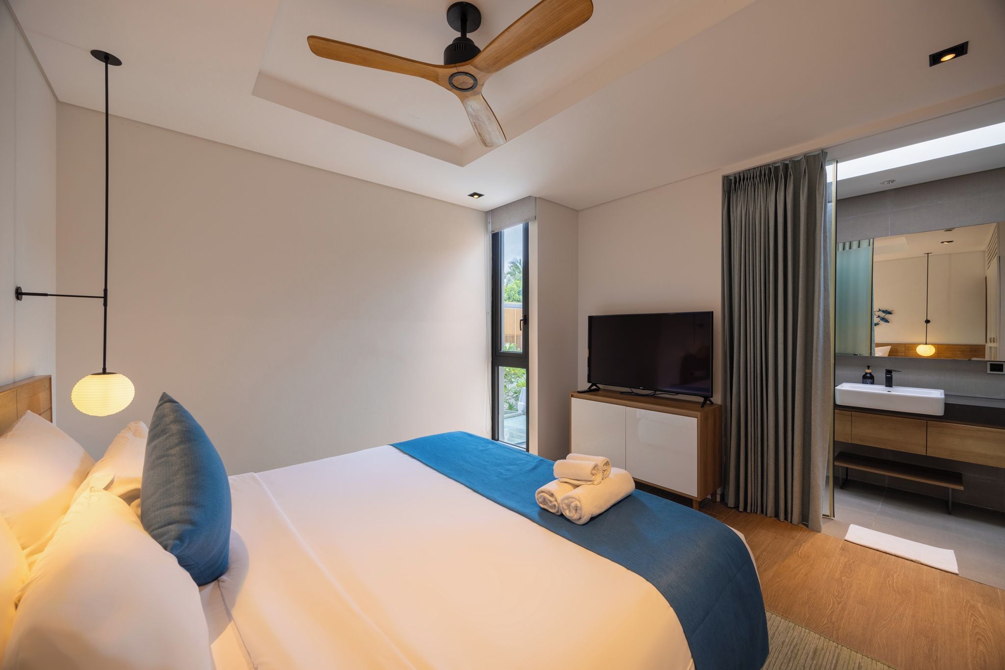 Una habitación de hotel moderna con una cama grande, un televisor de pantalla plana, un ventilador de techo de madera y un baño con espejo y lavamanos.