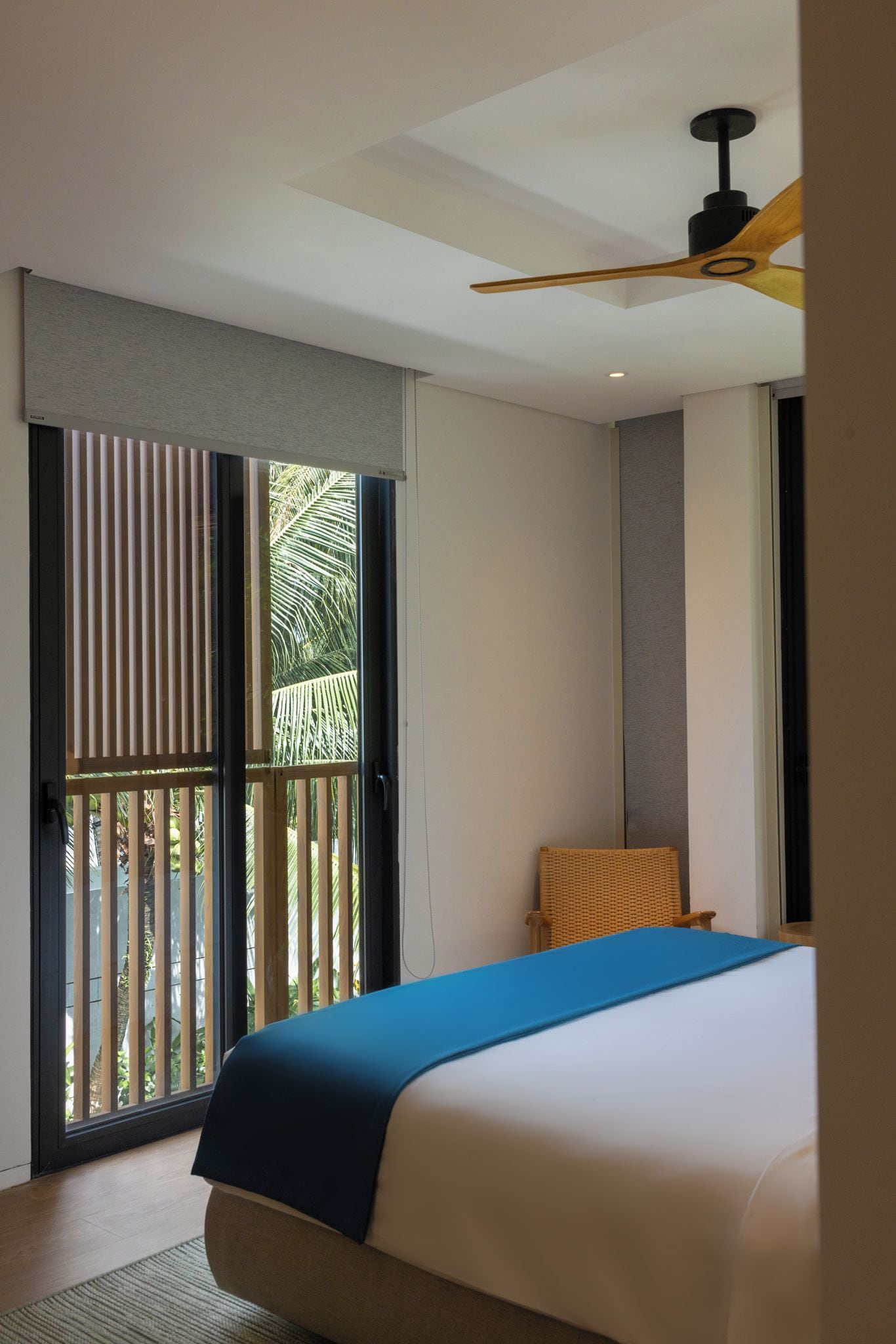 A hotel room with a bed, a ceiling fan, a window with blinds, and a view of palm trees.