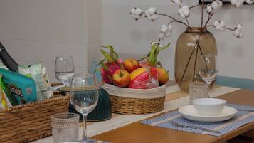 Una mesa de comedor con una cesta de frutas, copas de vino y un jarrón con plantas de algodón.