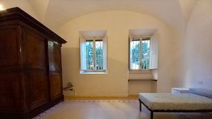 Una habitación con un amplio armario de madera, dos ventanas con marcos blancos, un tapete estampado y una cama con cubrecama gris.