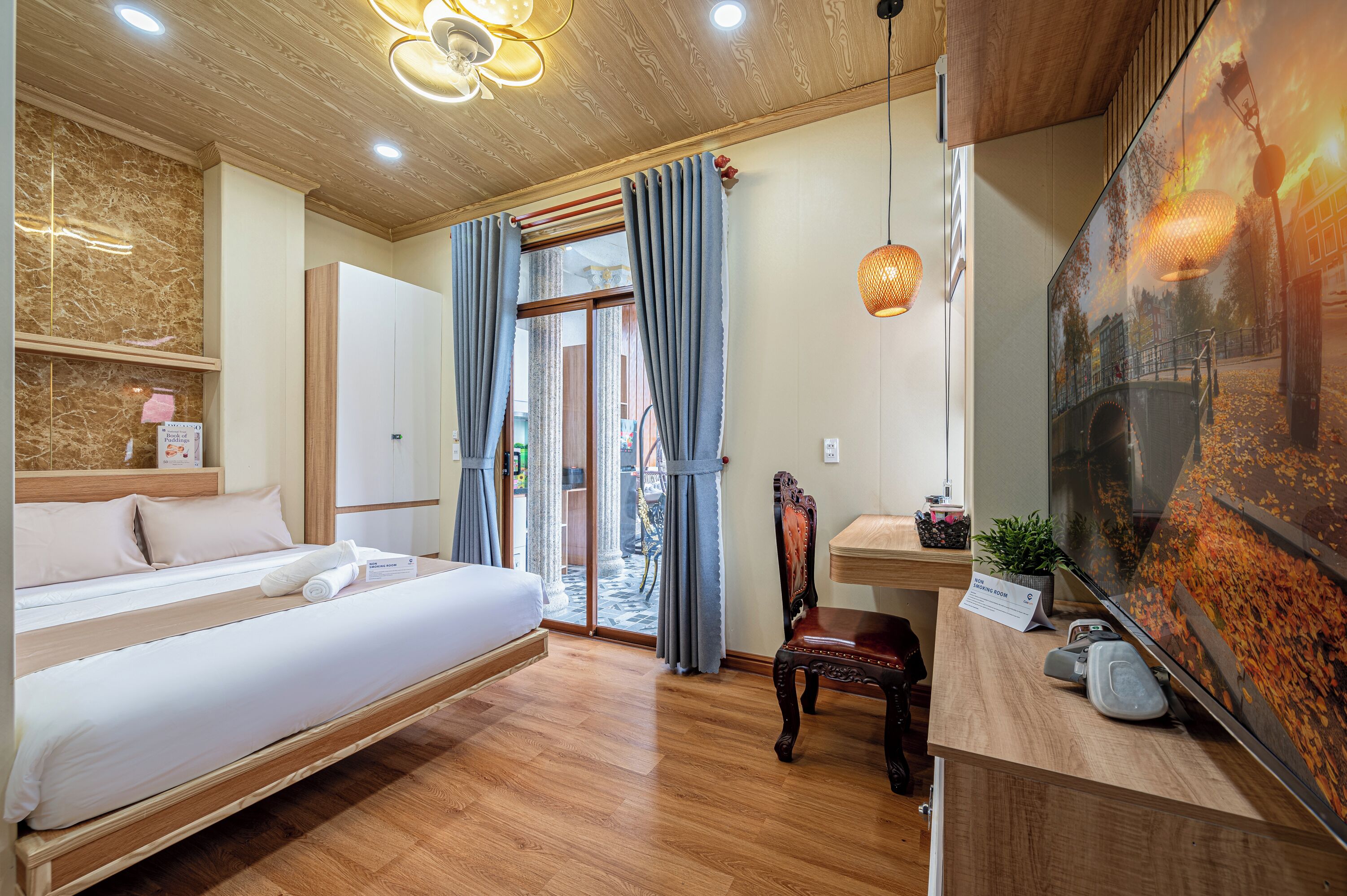 Ein Hotelzimmer mit einem Bett, einem Schreibtisch, einem Stuhl, einem großen Fenster mit Vorhängen und einem an der Wand befestigten Fernseher.