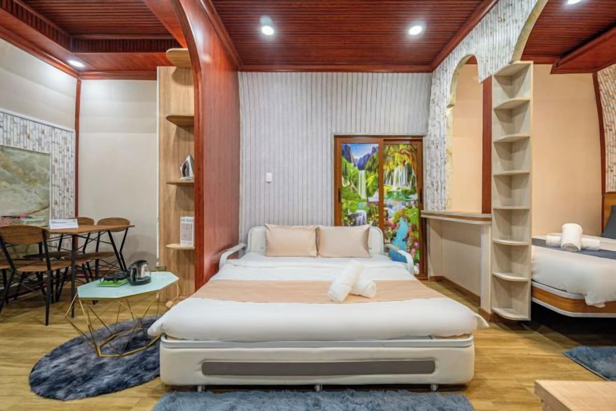 A hotel room with a bed, a desk, and a chair. There is a painting on the wall and a window with a view of greenery.