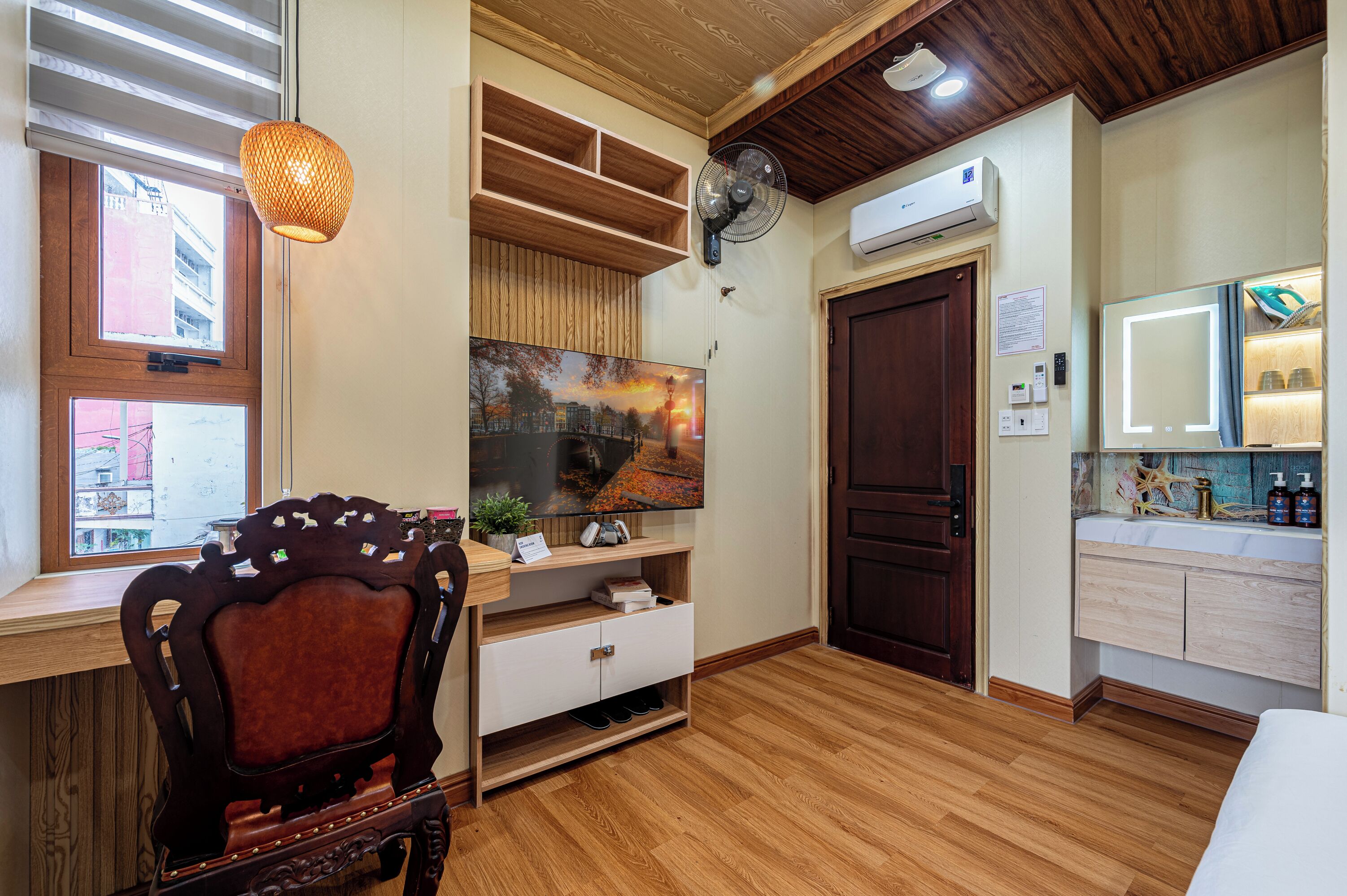 Una habitación con piso de madera, un televisor montado en un estante de madera, un ventilador, una puerta y una ventana con vistas a edificios.