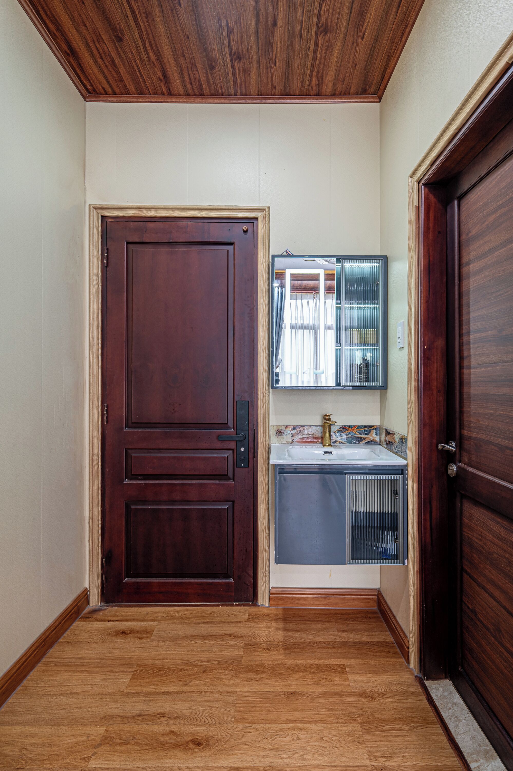 A hallway with a wooden door, a small bathroom with a mirror and sink, and a wooden floor.