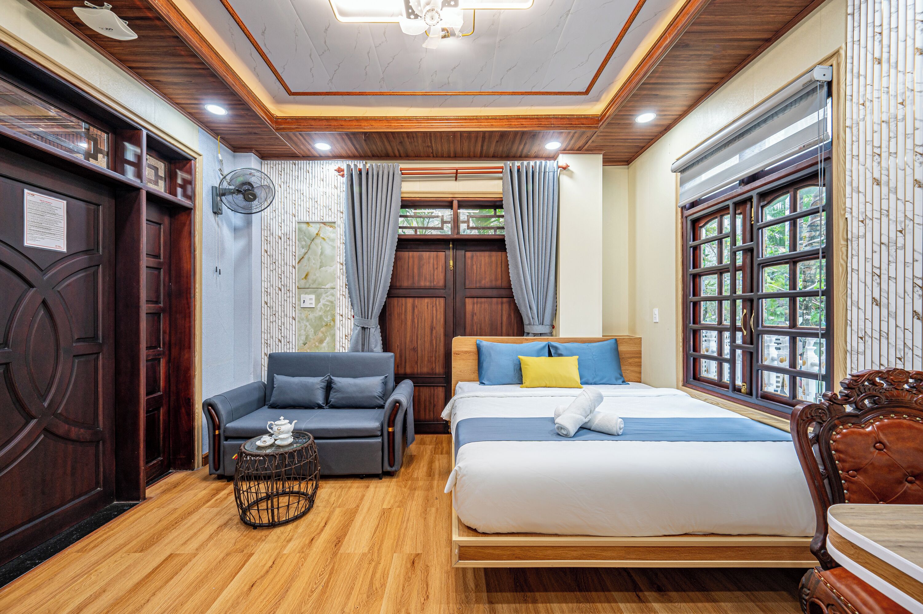 A hotel room with a wooden floor, a bed with blue and yellow bedding, a grey sofa, a small round table with a teapot, a wall-mounted fan, and a large window with a view of greenery.