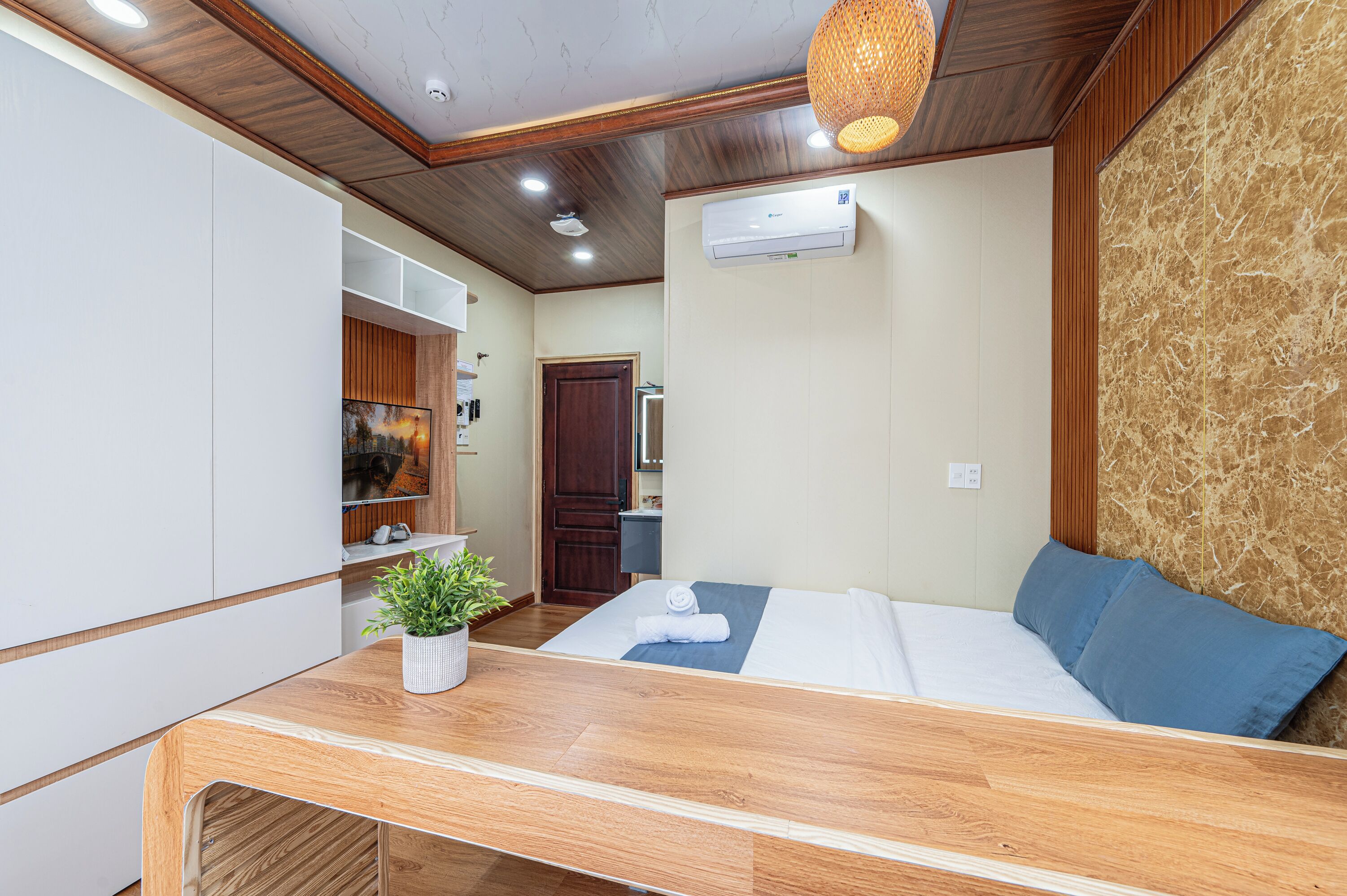 A hotel room with a wooden ceiling, a bed with blue pillows, a small table with a plant, and a wardrobe.