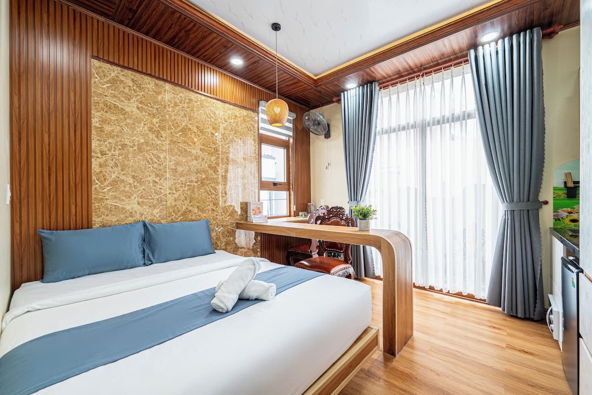 A modern hotel room with a large bed, wooden paneling, a marble accent wall, a small table with a fan, and a view of the outdoors through large windows.