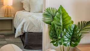 A modern bedroom with a bed, bedside table, lamp, and a large green plant. - Beautiful Marina View Apartment (Geraldton)