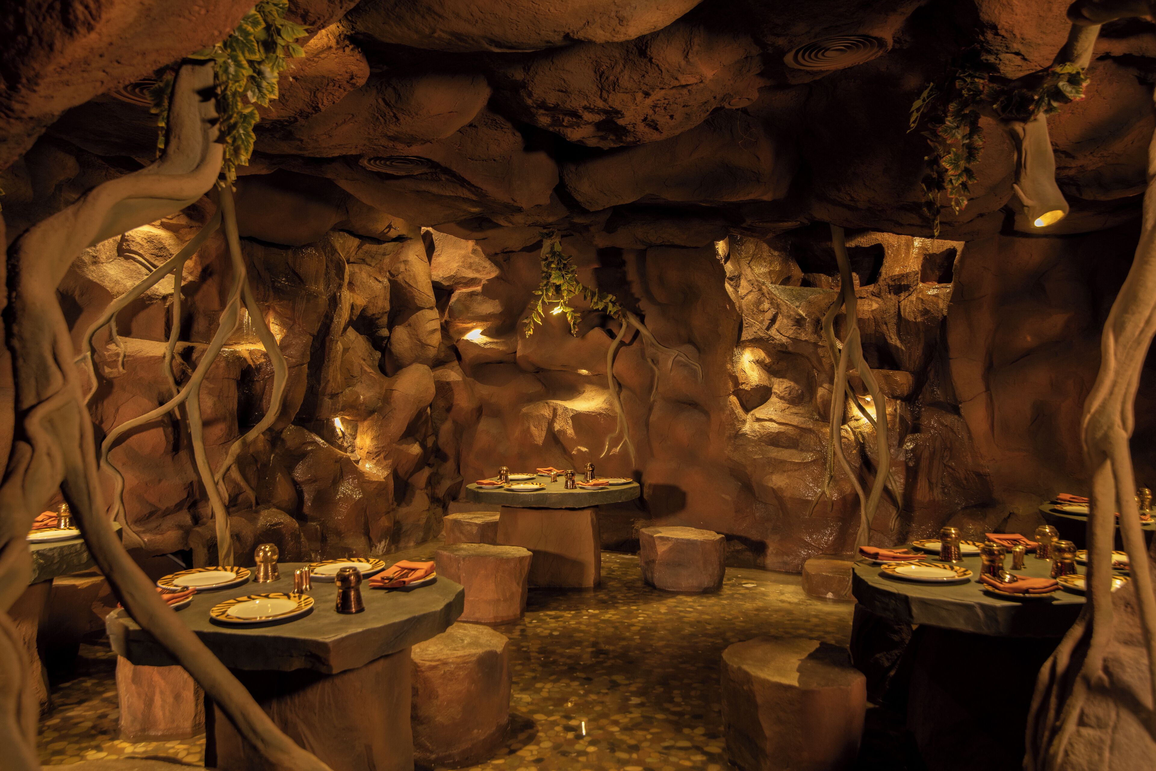 An underground dining area with tables set for a meal, surrounded by rock formations and hanging vines.