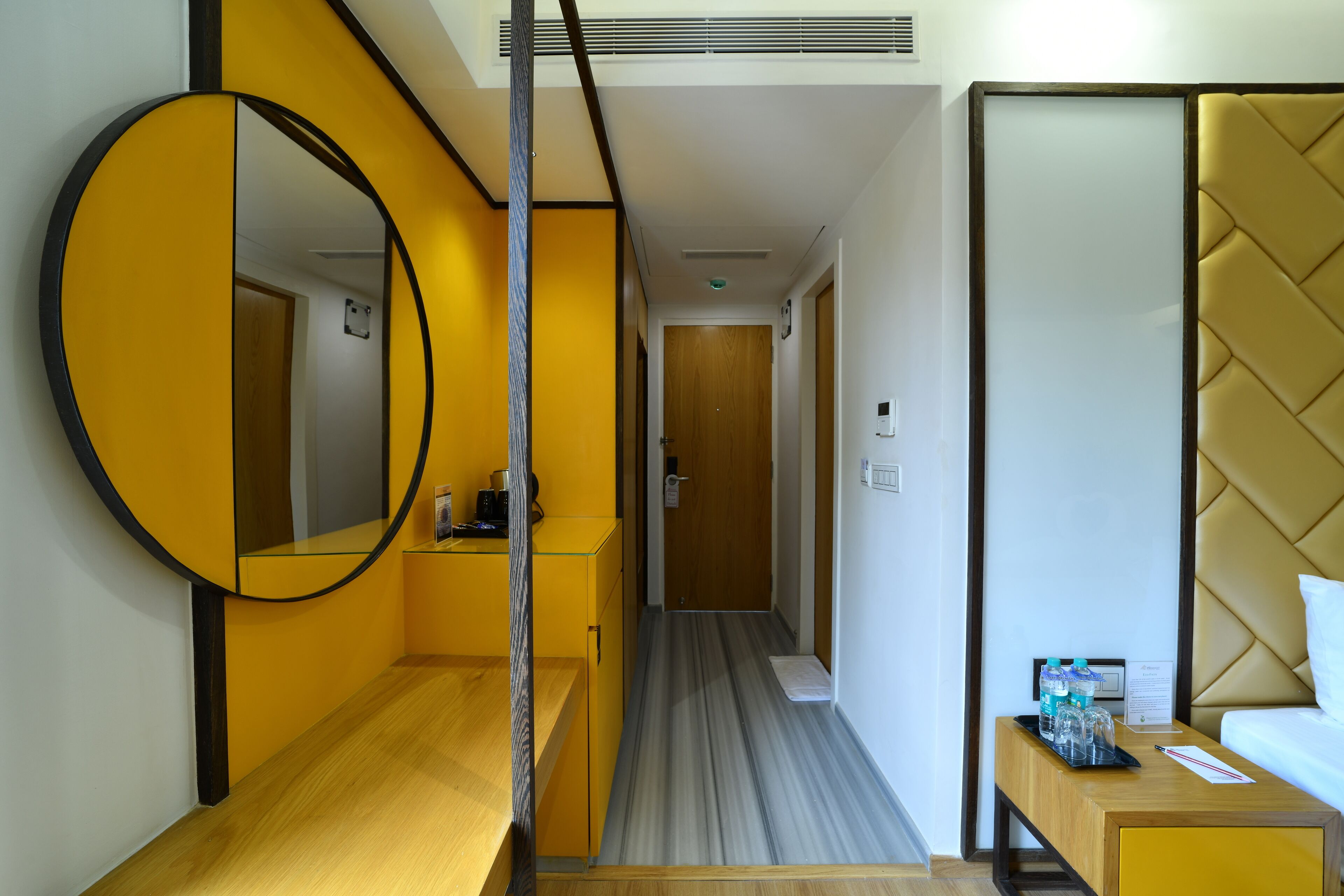 A hotel room with a wooden floor, a round mirror, a bed with a headboard, a bedside table with water bottles, and a wooden bench.
