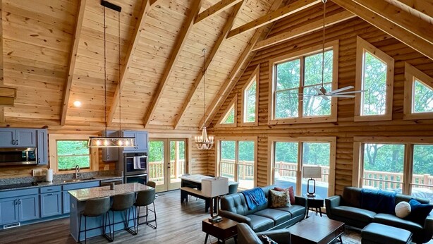 Un intérieur spacieux de chalet en bois, avec un plafond haut, un coin cuisine et un salon doté de sièges et d’une cheminée.