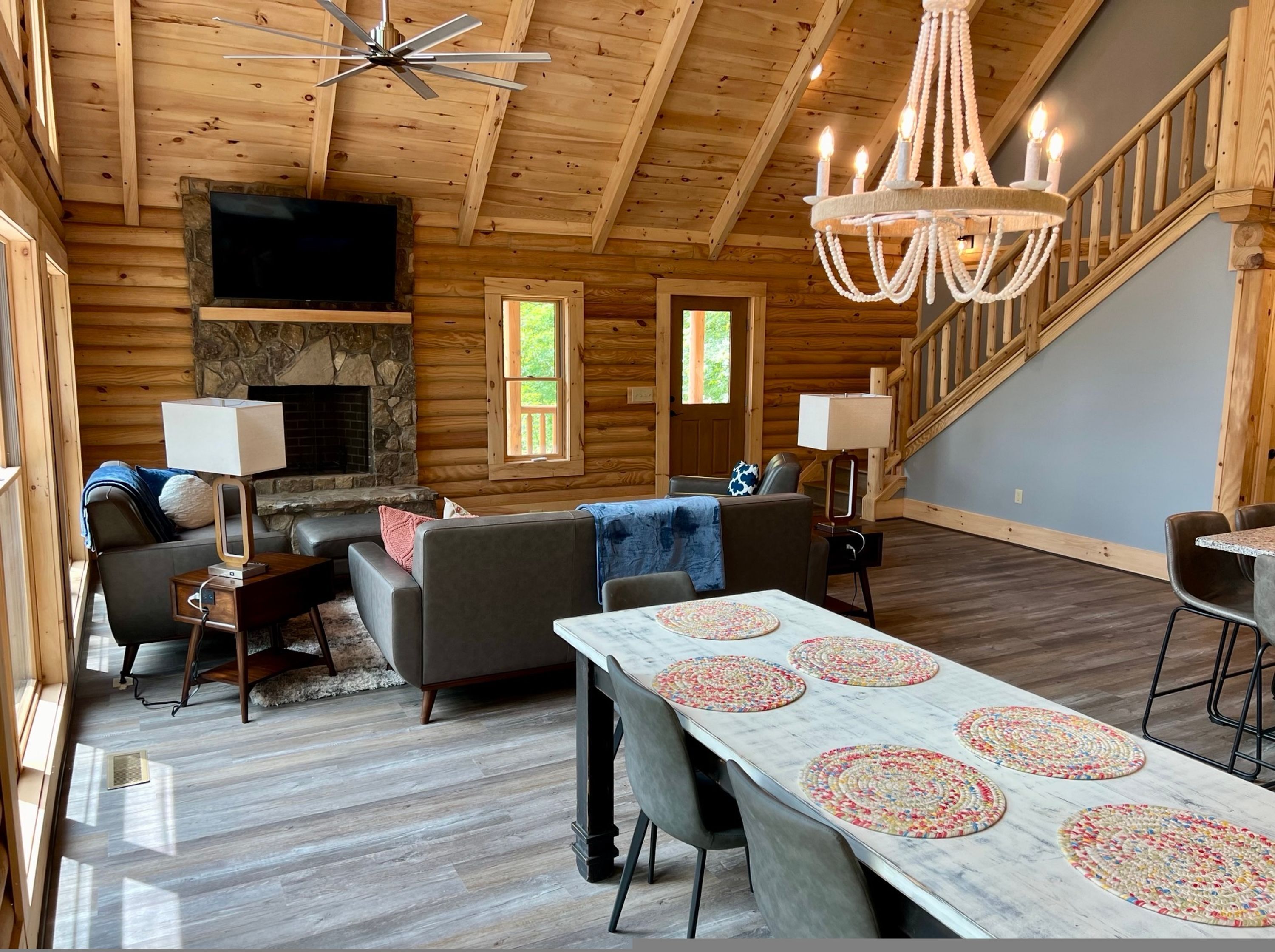 A log cabin interior with a stone fireplace, a dining table with placemats, a sofa set, a television, and a chandelier.