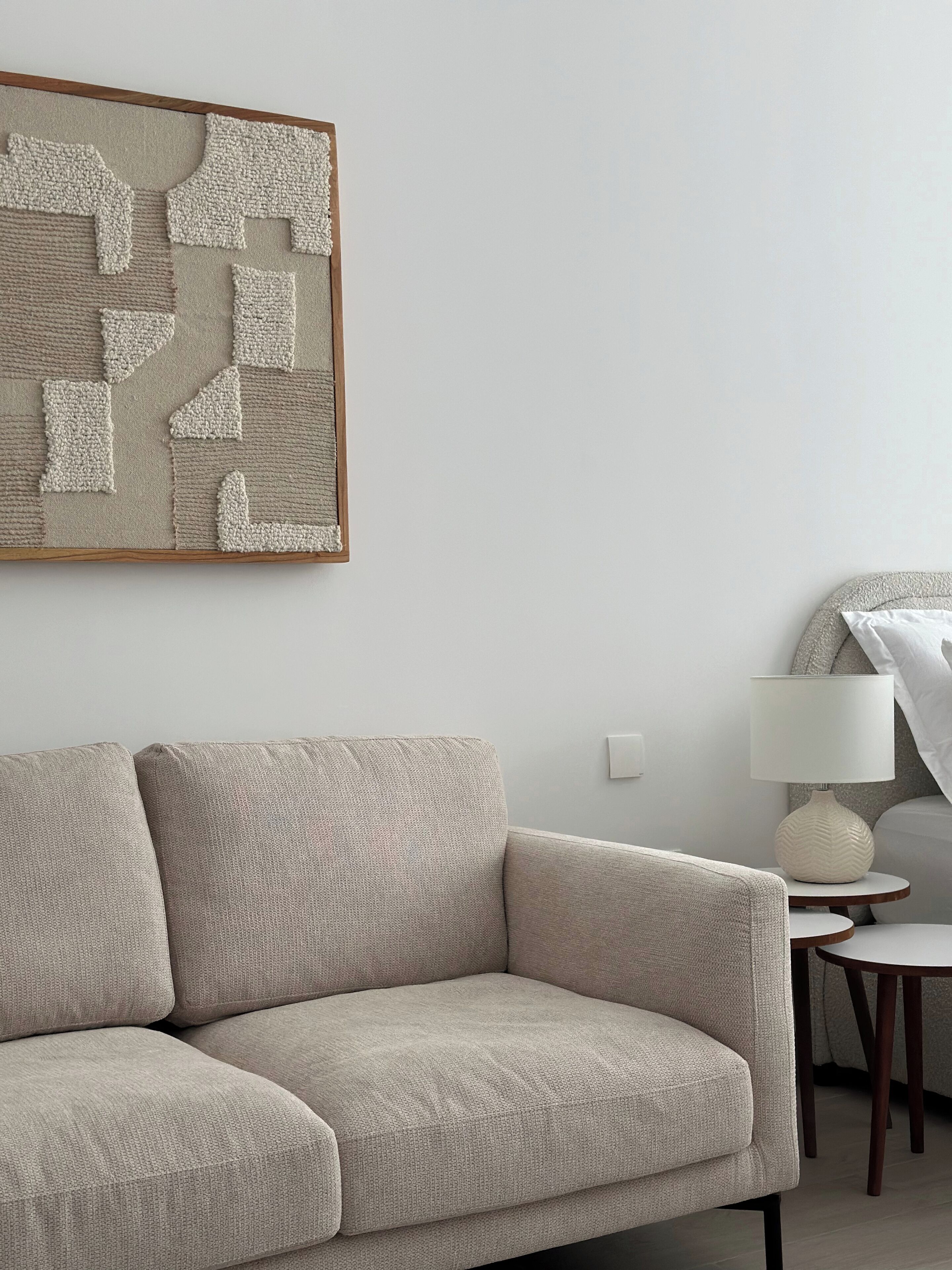 Ein modernes Wohnzimmer mit einem beigefarbenen Ecksofa, einem runden Holztisch, einer weißen Stehlampe und einem strukturierten Wandbild.