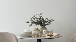 Une table de salle à manger ronde, ornée d’un vase rempli de verdure, de assiettes blanches et de tasses. Deux chaises aux pieds noirs sont placées de part et d’autre de la table.