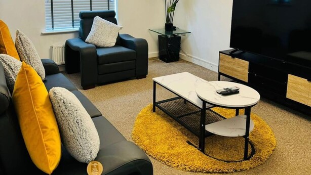 A living room with a black sofa, yellow cushions, a glass coffee table, and a TV. - House - Sleeps 8 - Parking - Garden (Peterborough)