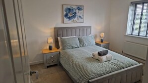 A bedroom with a bed, two bedside tables, a nightstand, a radiator, a window with blinds, and a wall-mounted painting. - House - Sleeps 8 - Parking - Garden (Peterborough)