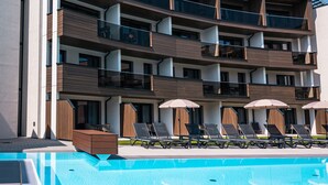 A modern multi-story hotel with a pool area, featuring balconies, lounge chairs, and umbrellas.