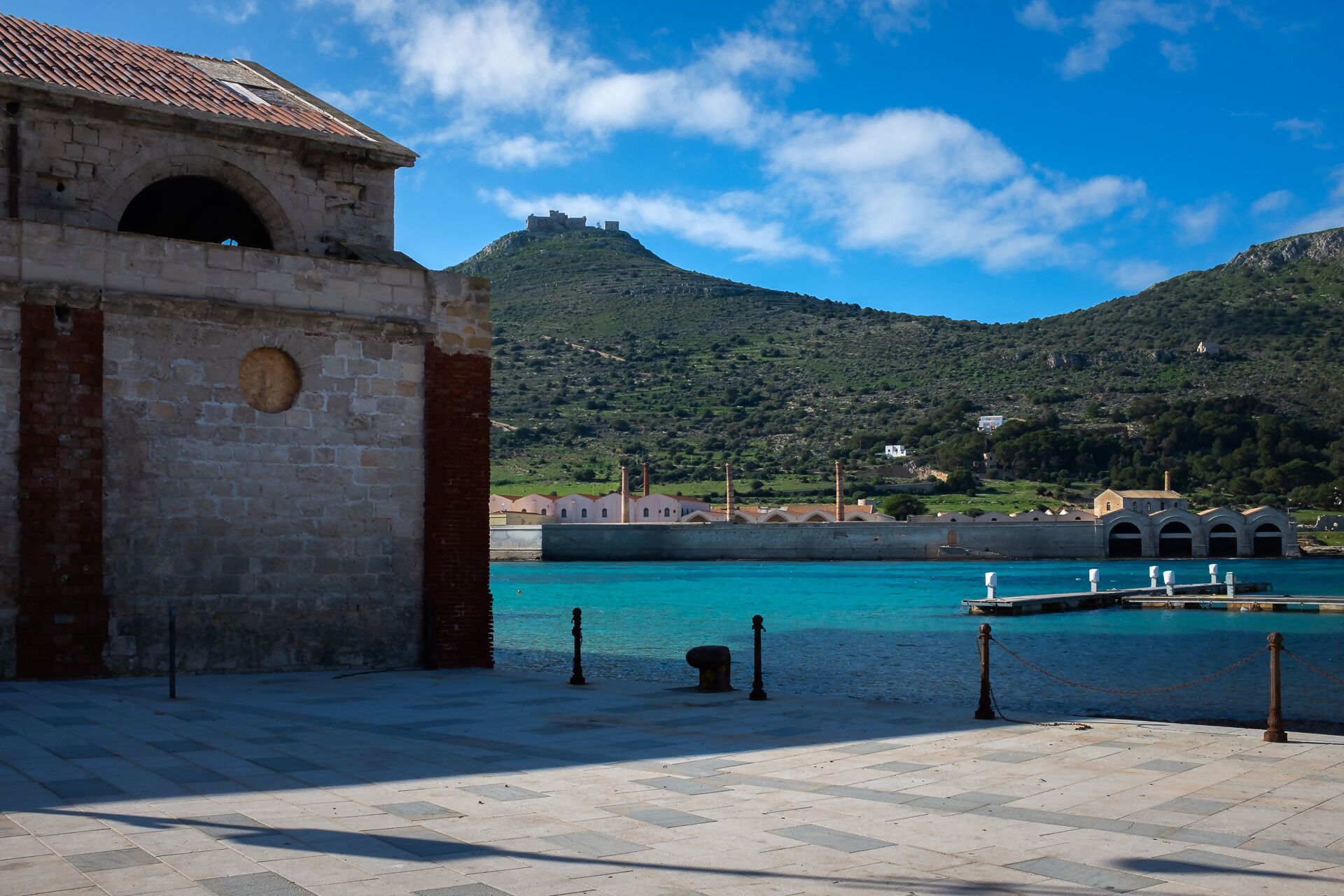 Un bâtiment situé sur la côte, doté d’une façade en pierre, d’une allée pavée et offrant une vue sur une mer turquoise et une colline en arrière-plan.