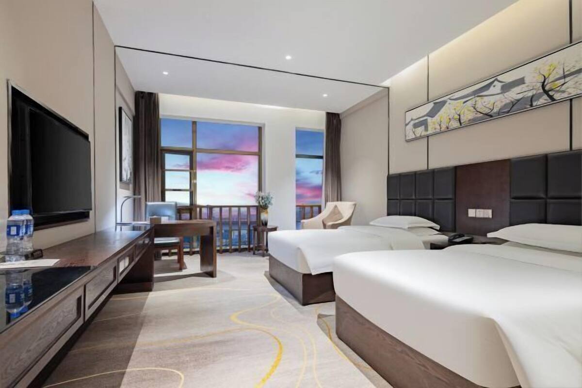 Une chambre d’hôtel avec un lit, un bureau doté d’une télévision, une chaise et une vue sur le ciel grâce à de grandes fenêtres.