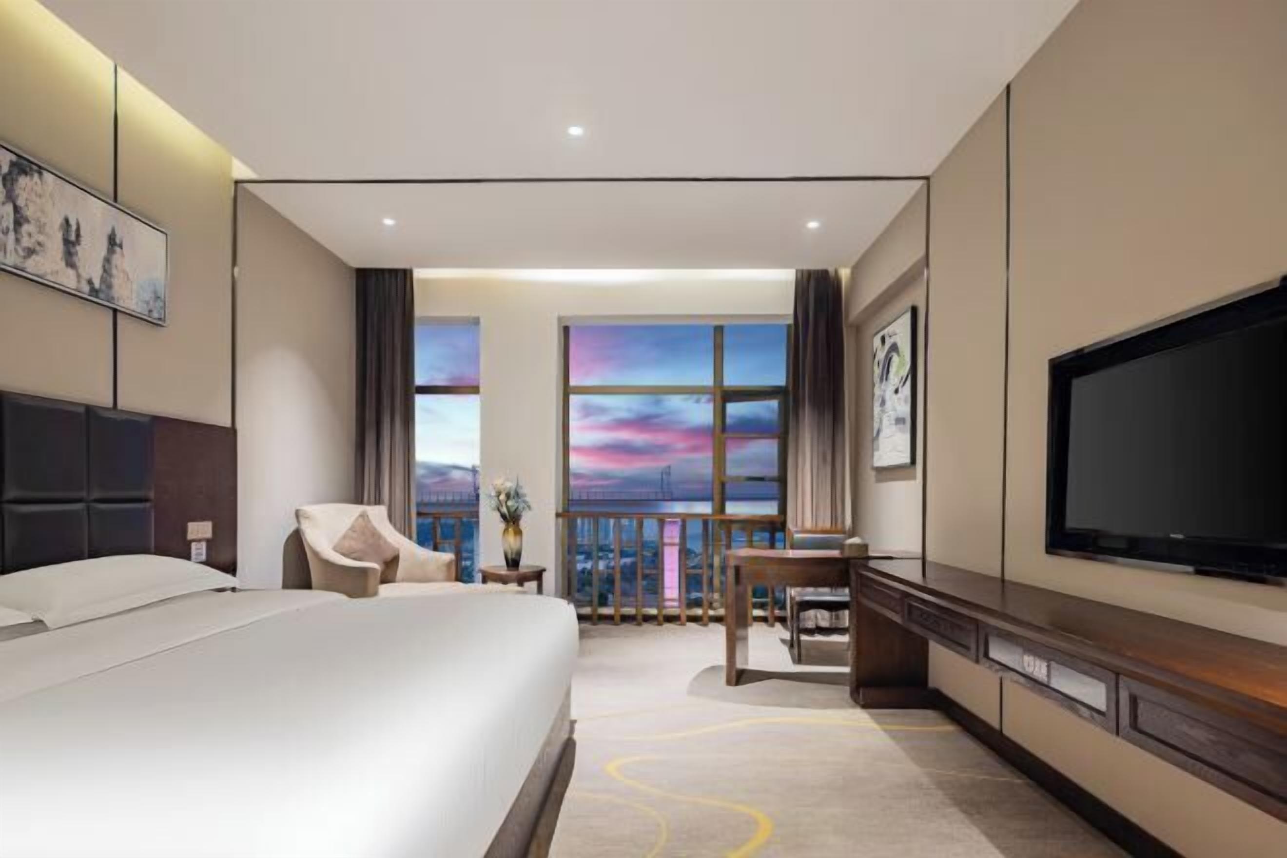 Habitación de hotel con cama, escritorio, silla, televisor y grandes ventanas con vista panorámica de la ciudad.