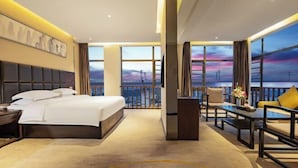Quarto de hotel com uma cama grande, uma escrivaninha, uma cadeira e vista para o pôr do sol através de grandes janelas.