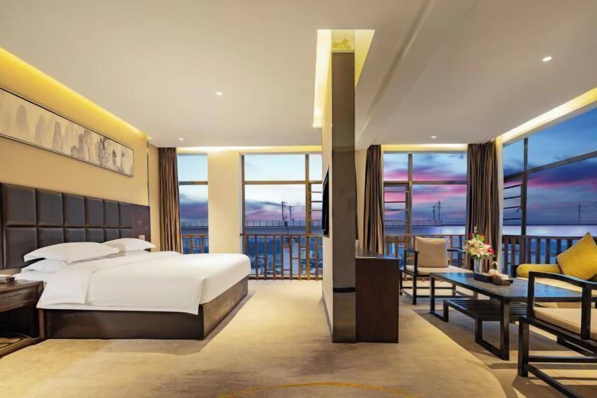 Une chambre d’hôtel avec un grand lit, un bureau, une chaise et une vue sur le coucher de soleil, visible à travers de grandes fenêtres.