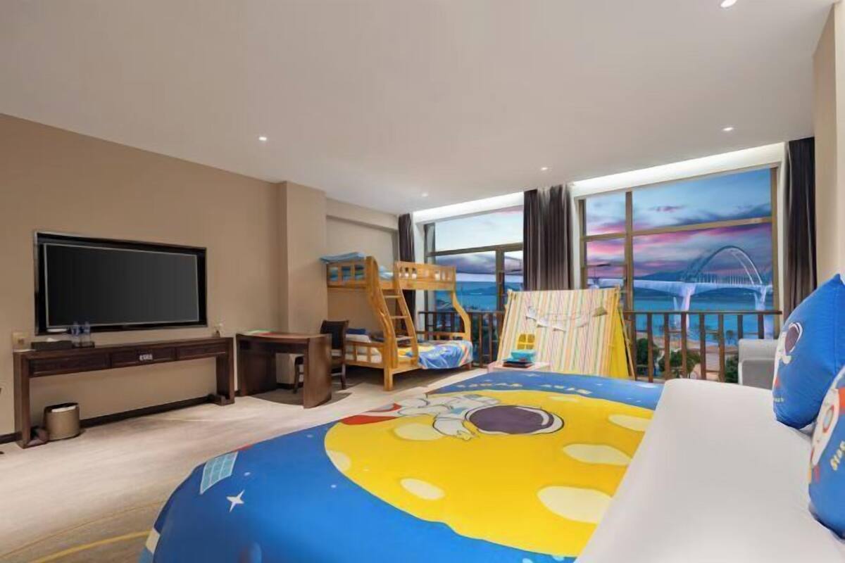Une chambre d’hôtel équipée d’un lit, d’un bureau, d’une télévision, d’un lit superposé et d’une grande fenêtre donnant sur le paysage urbain.