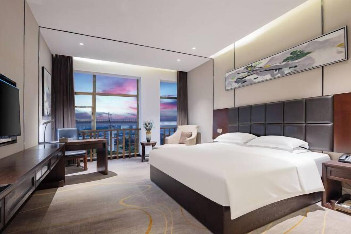 Une chambre d’hôtel moderne, dotée d’un grand lit, d’un bureau, d’une chaise, d’une télévision et offrant une vue sur le ciel par les fenêtres.