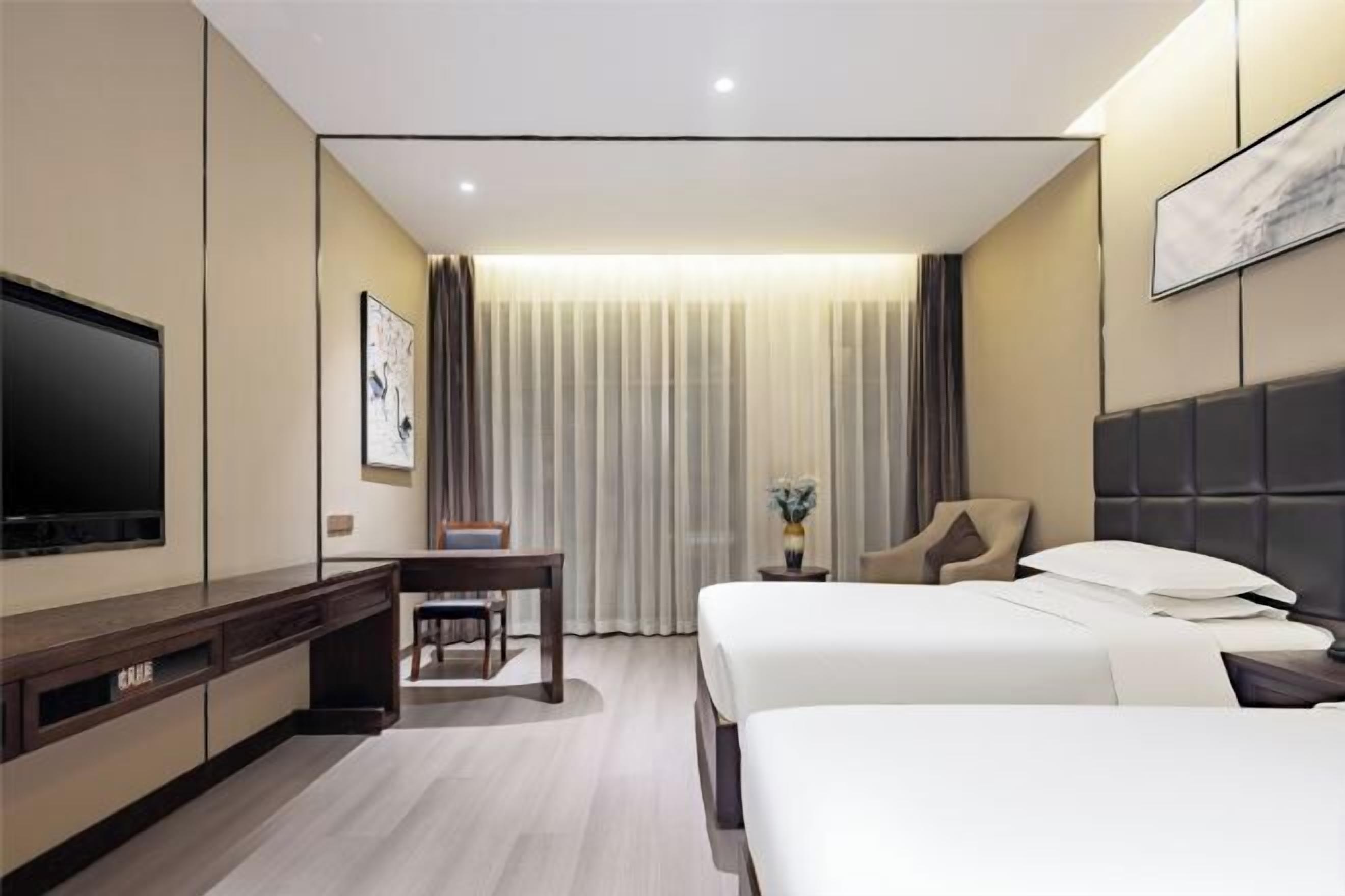 Ein Hotelzimmer mit zwei Betten, einem Schreibtisch, einem Stuhl, einem Fernseher und einem großen Fenster mit Vorhängen.