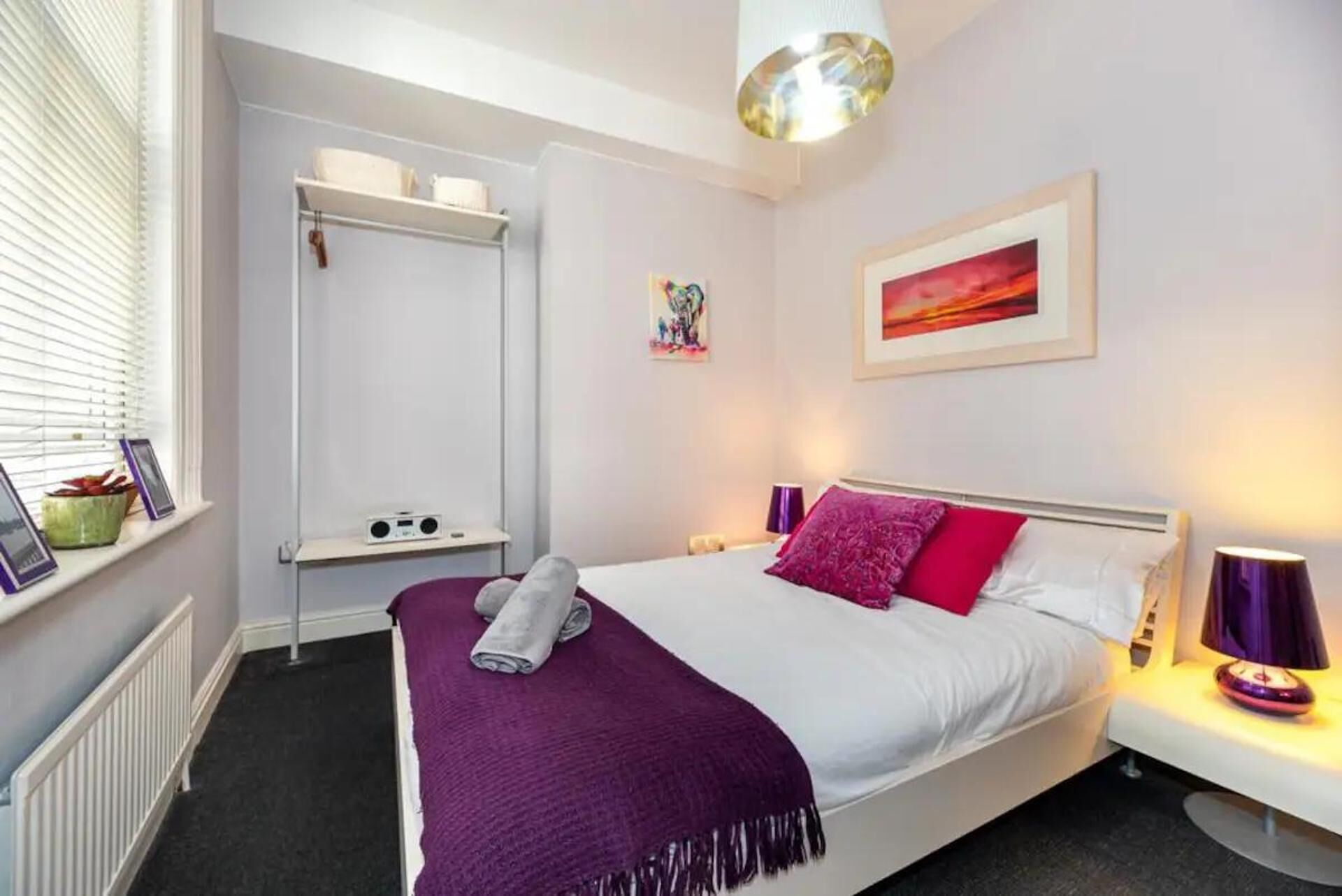 Ein Schlafzimmer mit Bett, Nachttisch, Lampe, einem Fenster mit Jalousien, einem Heizkörper und einem Regal mit Radio und Pflanze.