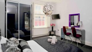 A modern bedroom with a bed, a desk, two red chairs, a TV, and a large mirror. - Designer Delight By Covent Garden Station (London)