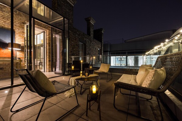 An outdoor seating area with wicker chairs and tables, a glass door entrance, and a view of the cityscape at night. - Penthouse Luxury in Covent Garden (London)