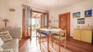 Un salon chaleureux avec un canapé, une table à manger et des chaises, un meuble en bois et une vue sur l’extérieur par une porte ouverte.