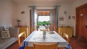Un coin repas avec une table dressée pour deux personnes, un canapé et une vue sur un jardin, visible à travers une porte ouverte.