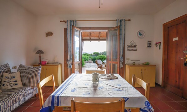Un coin repas avec une table dressée pour deux personnes, un canapé et une vue sur un jardin, visible à travers une porte ouverte.