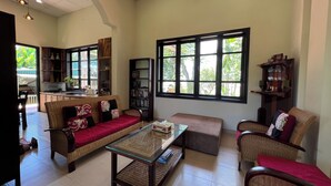 A spacious living area with a sofa, a coffee table, and a bookshelf. There are large windows providing natural light and a view of greenery outside.