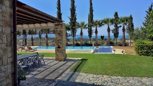 A stone patio with outdoor seating, a pool, and tall trees in the background. - Incredible Large Villa, Paphos, 6 bedrms BBQ Huge Pool Childrens pool Quiet area (2CQW+J39 Poli Crysochous, Cyprus)