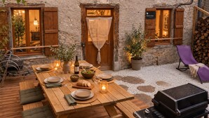 Un espace de restauration extérieur rustique, avec une table en bois dressée pour deux personnes, entouré de murs en pierre et de volets en bois.