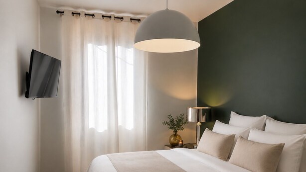 Une chambre d’hôtel avec un lit, un téléviseur fixé au mur, une table de chevet avec une lampe et une fenêtre avec des rideaux.