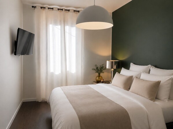 Habitación de hotel con cama, televisor montado en la pared, mesita de noche con lámpara y ventana con cortinas.
