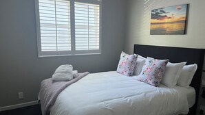 Una cama bien hecha con sábanas blancas, un cojín floral y una manta. Hay una ventana con persianas, un cuadro de un atardecer y un enchufe en la pared.