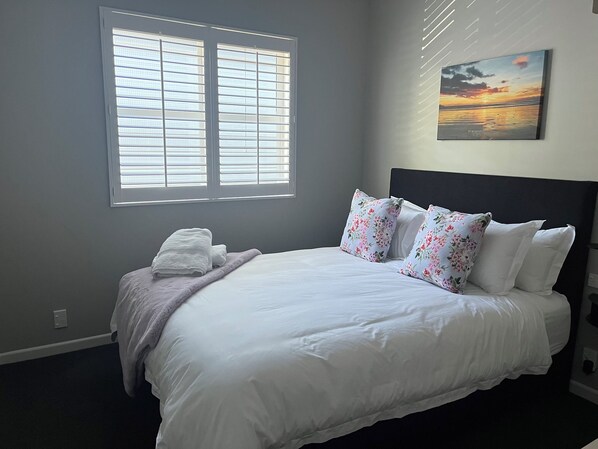 Una cama bien hecha con sábanas blancas, un cojín floral y una manta. Hay una ventana con persianas, un cuadro de un atardecer y un enchufe en la pared.