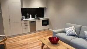 Una cocina moderna con armarios blancos, lavavajillas y horno. Hay un sofá gris con cojines estampados y una mesa de centro de madera con un tazón rojo.