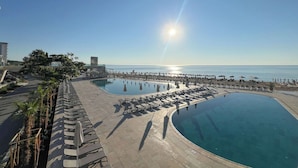 Zona de piscina en un resort con múltiples tumbonas y sombrillas, vista despejada al mar y un edificio grande al fondo.