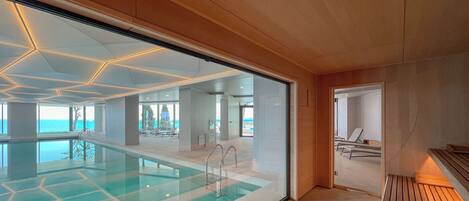 A modern indoor swimming pool with geometric ceiling lights, clear glass walls, and a view of the sea.