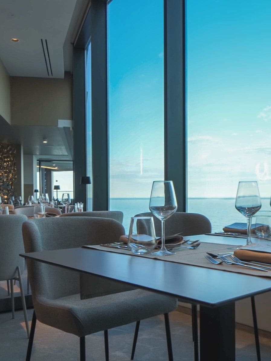 Un coin repas avec une table dressée pour deux personnes, de grandes fenêtres et une vue sur la mer.