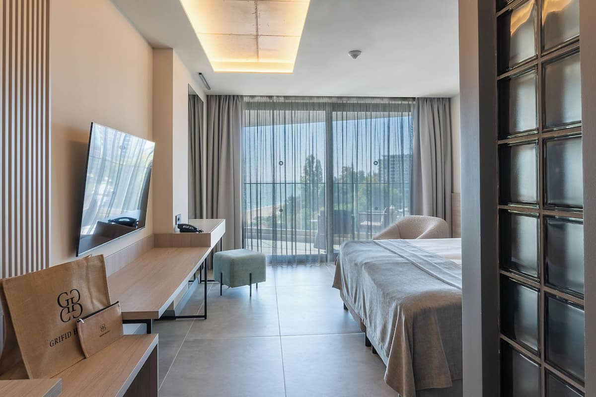 Una habitación de hotel moderna con cama, escritorio, silla, televisor y un ventanal con cortinas.