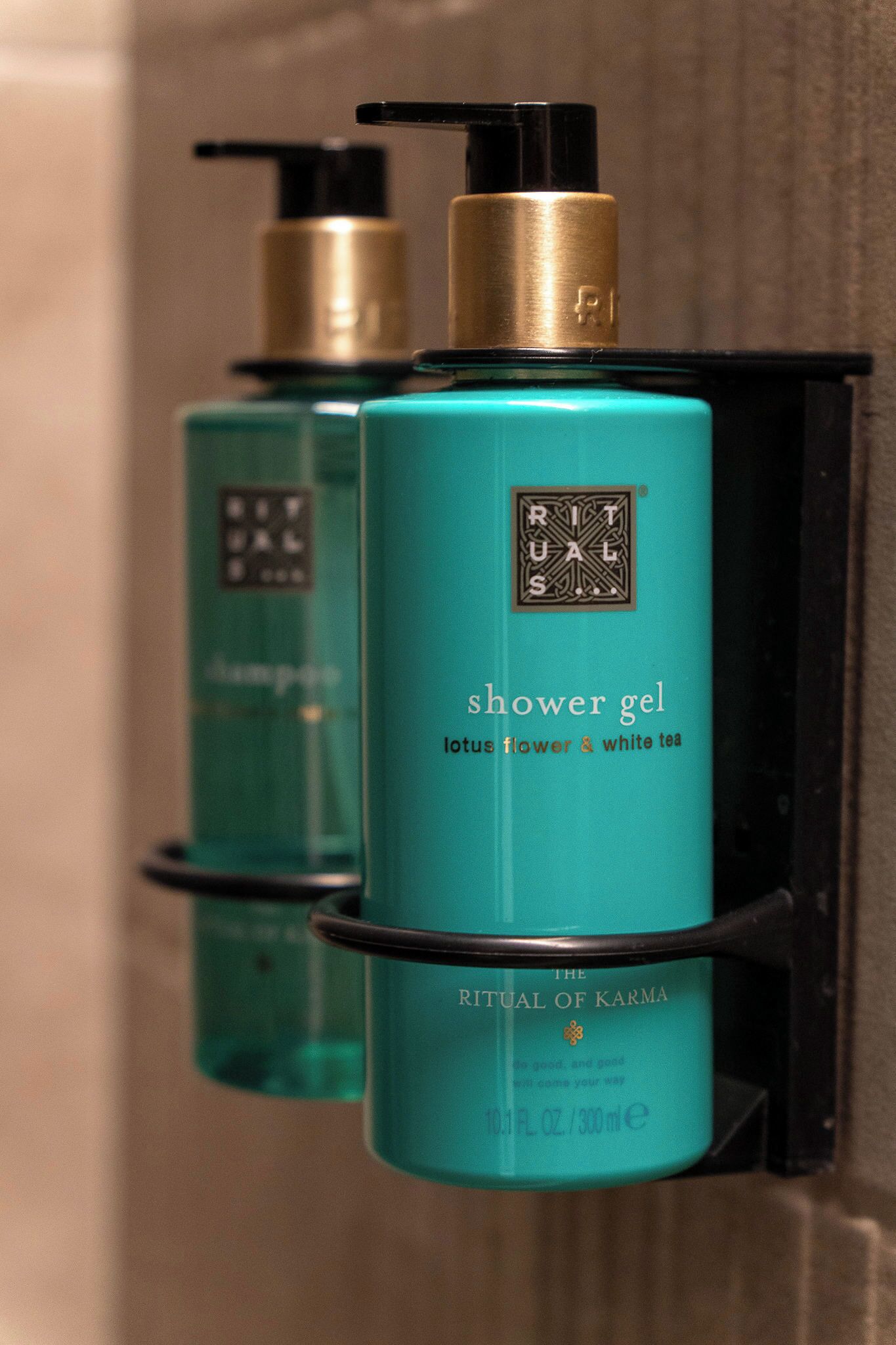 Deux flacons de gel douche avec un distributeur à pompe, portant les mentions « Rituals » et « The Ritual of Karma », sont posés sur un porte-savon noir fixé au mur.
