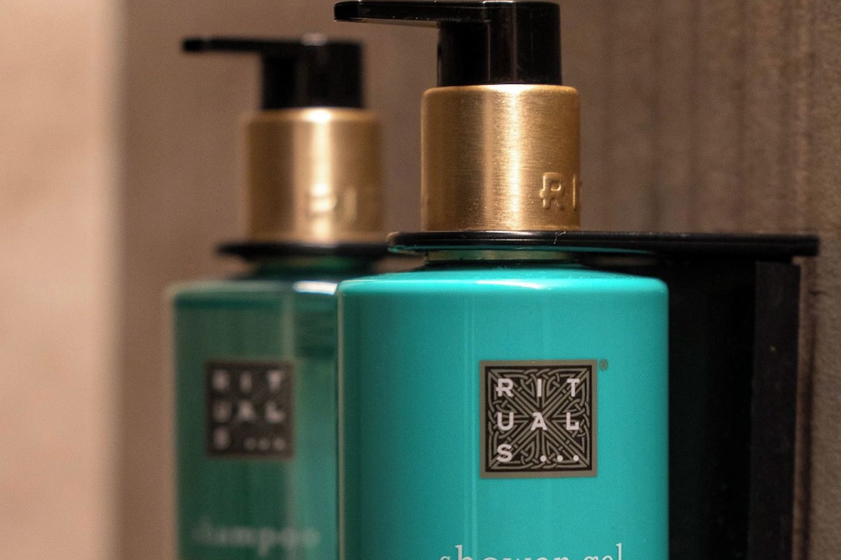 Dos botellas de gel de ducha con dosificador bomba, etiquetadas 'Rituals' y 'The Ritual of Karma', sobre un estante fijado a la pared negra.