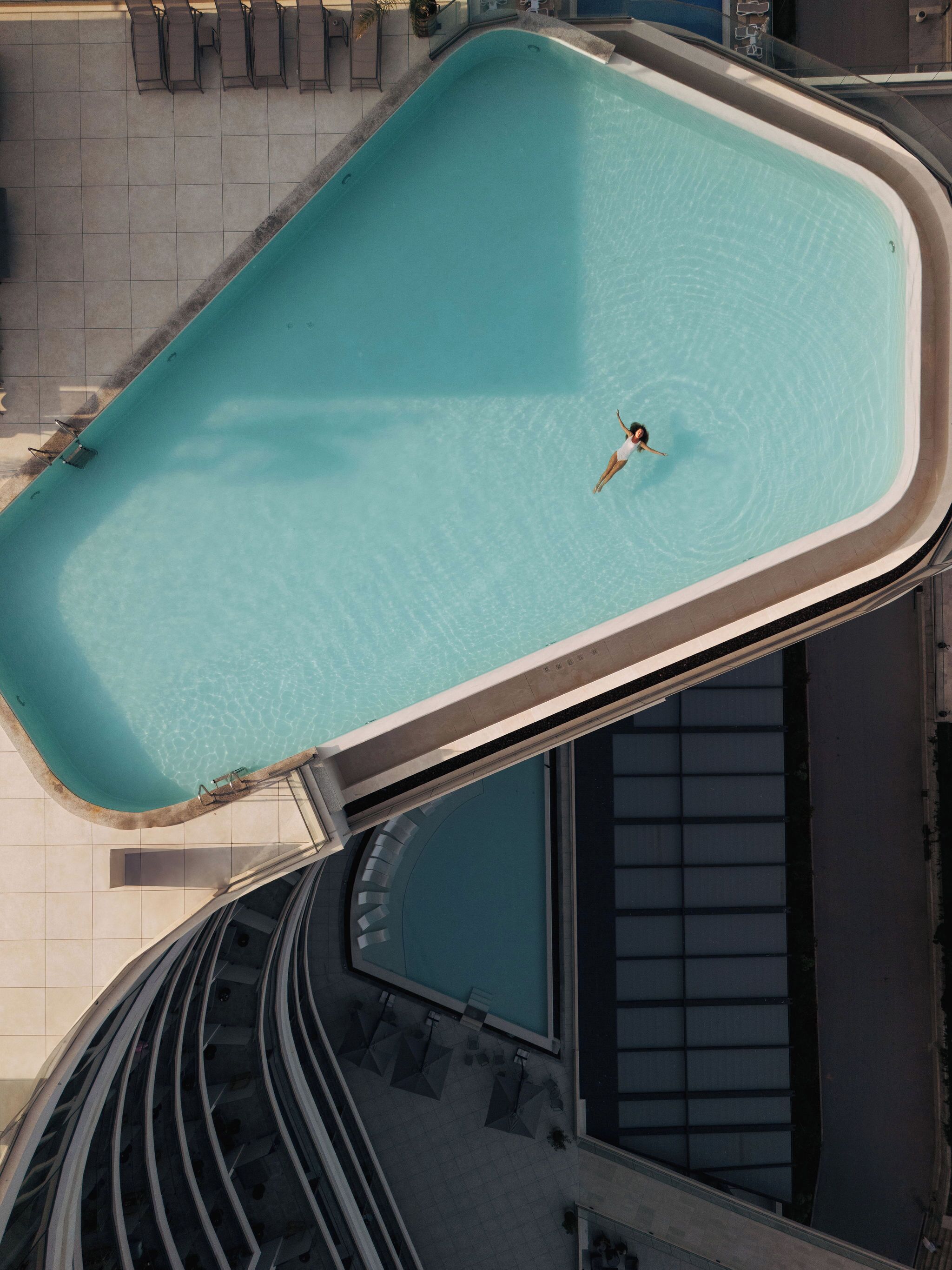 Vista aerea di una piscina con una persona che si tuffa, circondata da sdraio e un pavimento piastrellato.